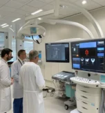 A photograph of a modern cardiology suite interior featuring a sleek, stainless steel cardiac monitor screen displaying heart rate data, a polished examination table with a patient lying supine, and a doctor in a crisp white lab coat adjusting the monitor, all illuminated by cool, diffused overhead lighting that highlights the clinical precision of the environment.